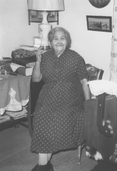 Historic photograph of Carrie Lodge in her home. She wears a pokadot dress and sits on a chair with a smile on her face.